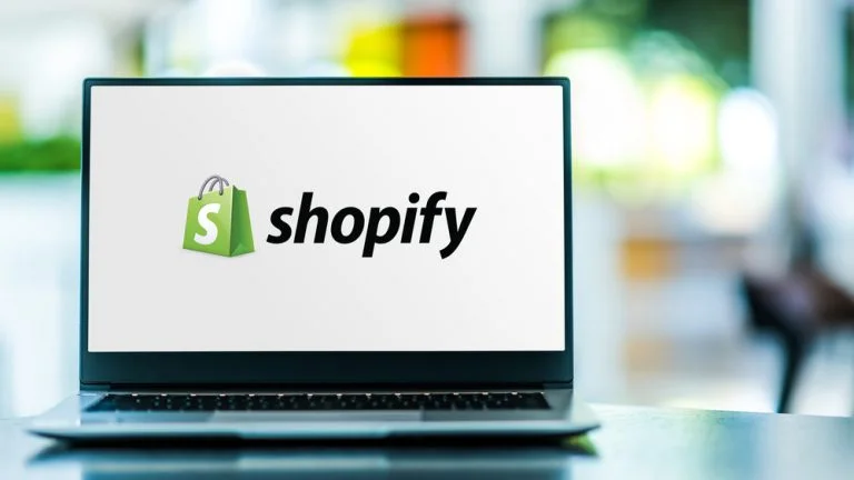 Laptop displaying the Shopify logo on its screen. The logo consists of a green shopping bag with a white 'S' on it, followed by the word 'shopify' in black text. The background of the image is blurred, suggesting an indoor setting.