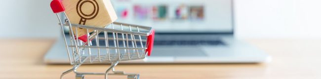 Shopping,Cart,With,Magnifying,Icon,Block,And,Laptop,Computer,With A miniature shopping cart with a red handle holding a wooden block featuring a magnifying glass icon. The cart is placed on a wooden surface in front of a blurred laptop screen, indicating an online shopping or e-commerce concept.
