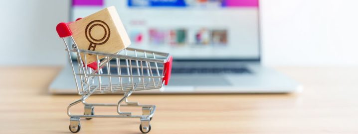 Shopping,Cart,With,Magnifying,Icon,Block,And,Laptop,Computer,With A miniature shopping cart with a red handle holding a wooden block featuring a magnifying glass icon. The cart is placed on a wooden surface in front of a blurred laptop screen, indicating an online shopping or e-commerce concept.
